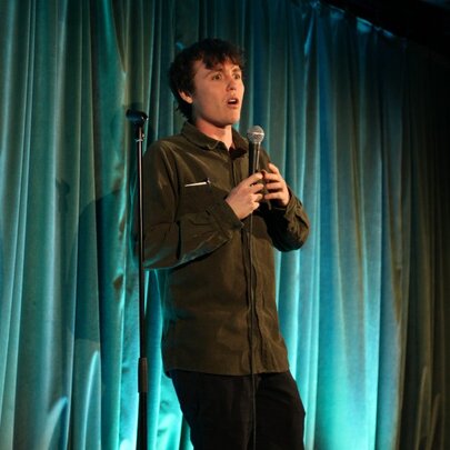 A stand-up comedian talking into a microphone they hold in their hand, performing live on stage.