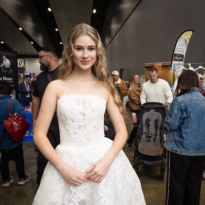 A young woman in a strapless bridal gown in an expo hall with people gathered in the background. 