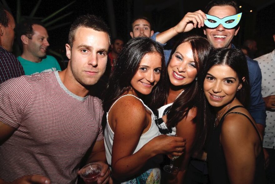 Five beautiful people posing for a photo at a nightclub, one holding a blue masquerade mask over his eyes.