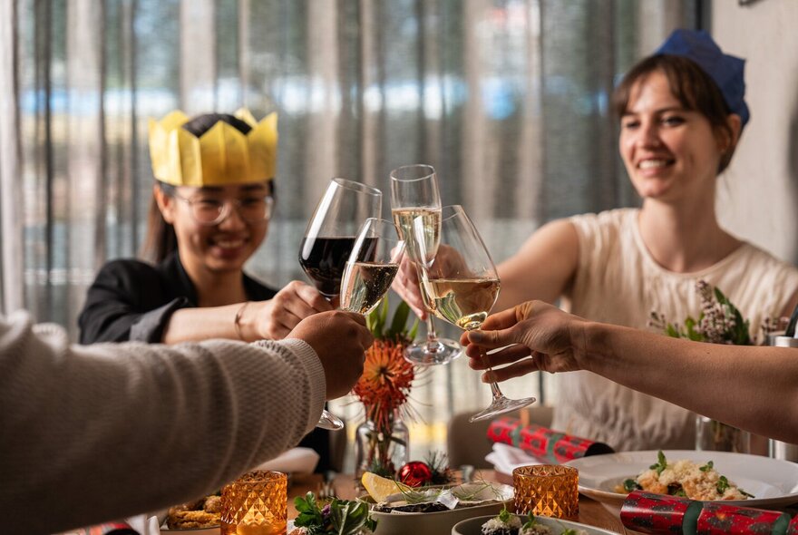 Friends sitting around a dining table in a restaurant, wearing Christmas hats, and bringing their glasses together to clink.