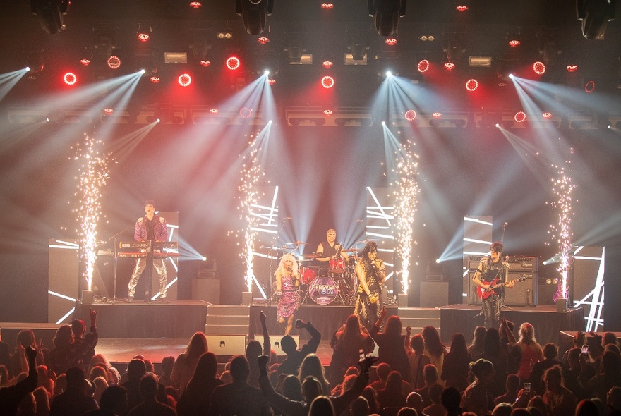 Five 80s-style rockers on stage with theatrical flames and strobe lighting, in front of a crowd.