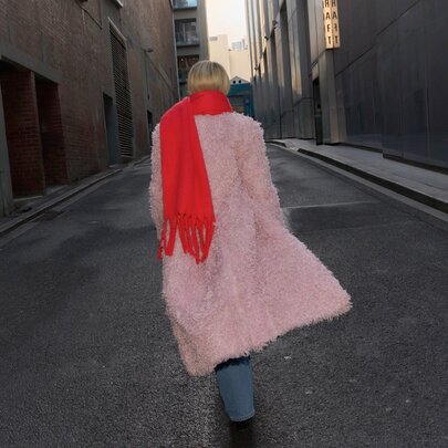 A woman is walking down an alley way wearing a pink coat and a red scarf