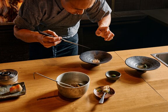 A Japanese chef bent over an earthenware dish featuring a single slice of daikon, chopsticks in hand.