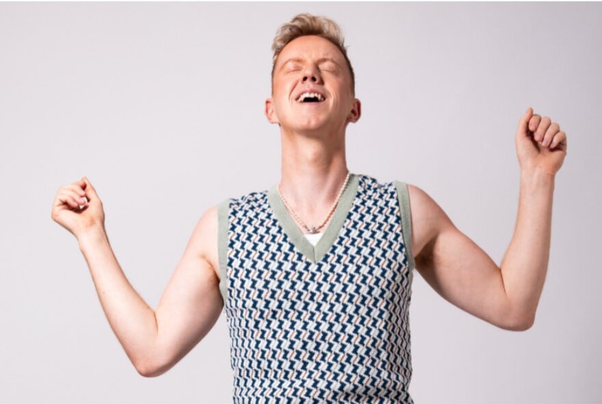 Young man with short blond hair wearing a knitted vest, his arms bent and pumped.