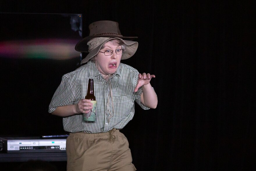 Comedian in character outfit dressed as an older man, wearing a hat and holding a bottle.