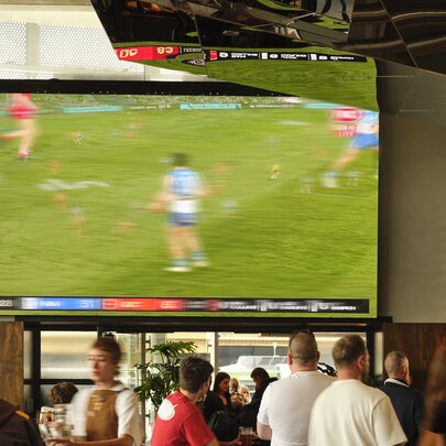 People walking around inside a bar with a large monitor on the wall screening a sporting match.