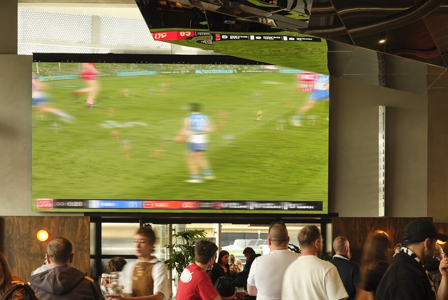 People walking around inside a bar with a large monitor on the wall screening a sporting match.