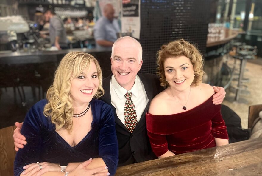 One male and two female opera singers dressed in formal attire, seated at a wooden table while posing for a photo. 