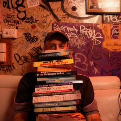 Person seated on a couch with graffiti scrawled on the walls, a pile of books on their lap.