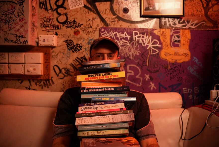 Person seated on a couch with graffiti scrawled on the walls, a pile of books on their lap.