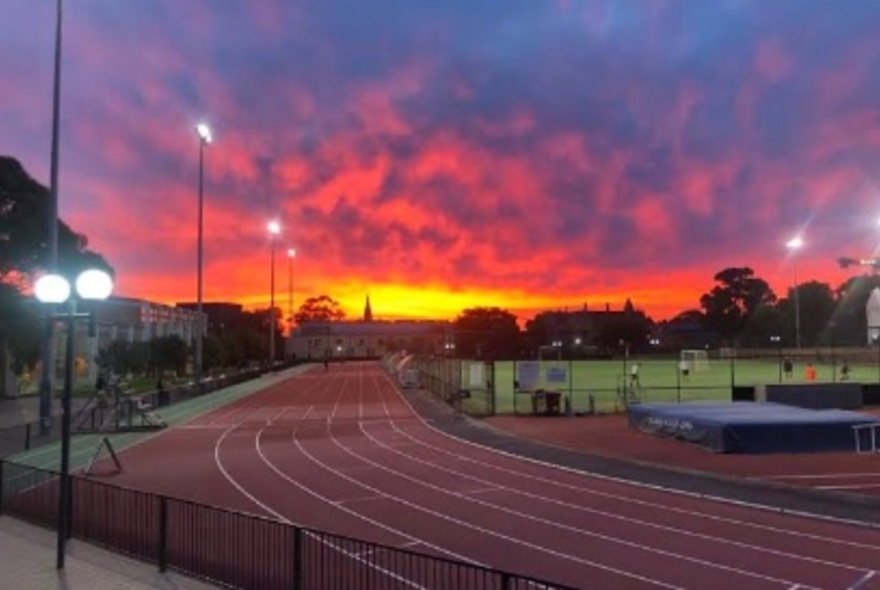 The curve of a red running track with a vibrant sunset behind and a soccer pitch on the right.