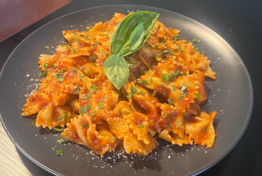 Bowtie pasta with a napoli sauce and two basil leaves on top, on a black plate.