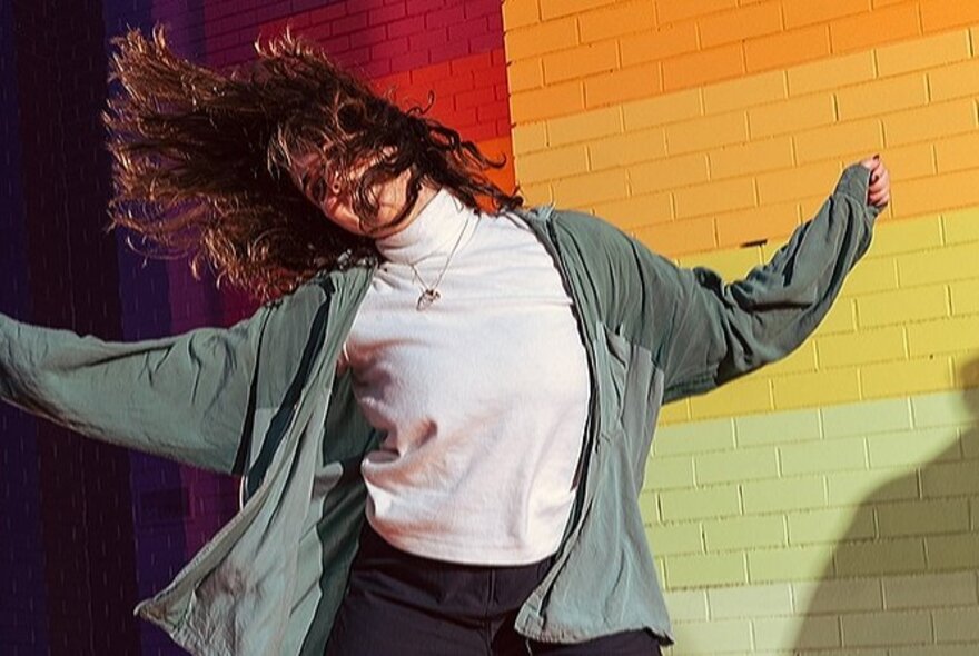 A woman dancing in front of a rainbow-coloured brick wall, her long brown hair fanning out around her.