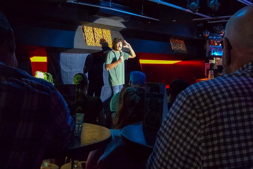 People seated in a dark bar venue watching a stand-up comic performing on stage, holding a microphone.