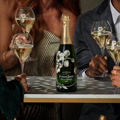 A bottle of Perrier-Jouët Belle Époque Champagne with its iconic hand-painted anemone design, surrounded by people holding champagne flutes.