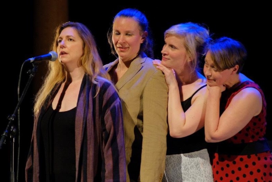 Three female improvisers standing in a line behind a fourth who speaks into a microphone on a stage. 