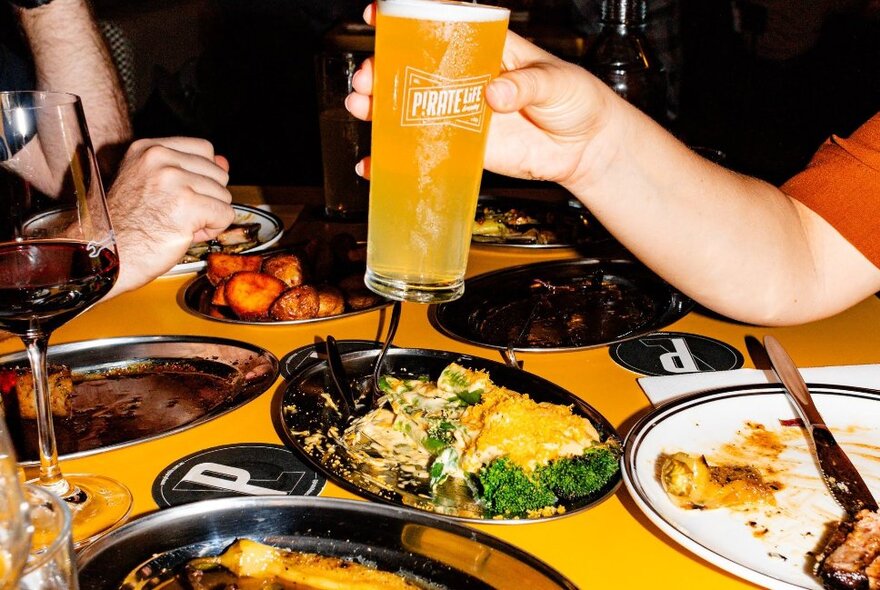 Hand holding a glass of beer over dishes of food on a restaurant rable.