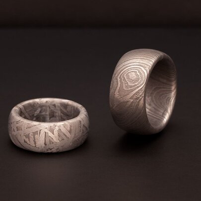 Two thick, patterned metal rings sit on a dark surface, with one lying flat and the other standing on its edge.
