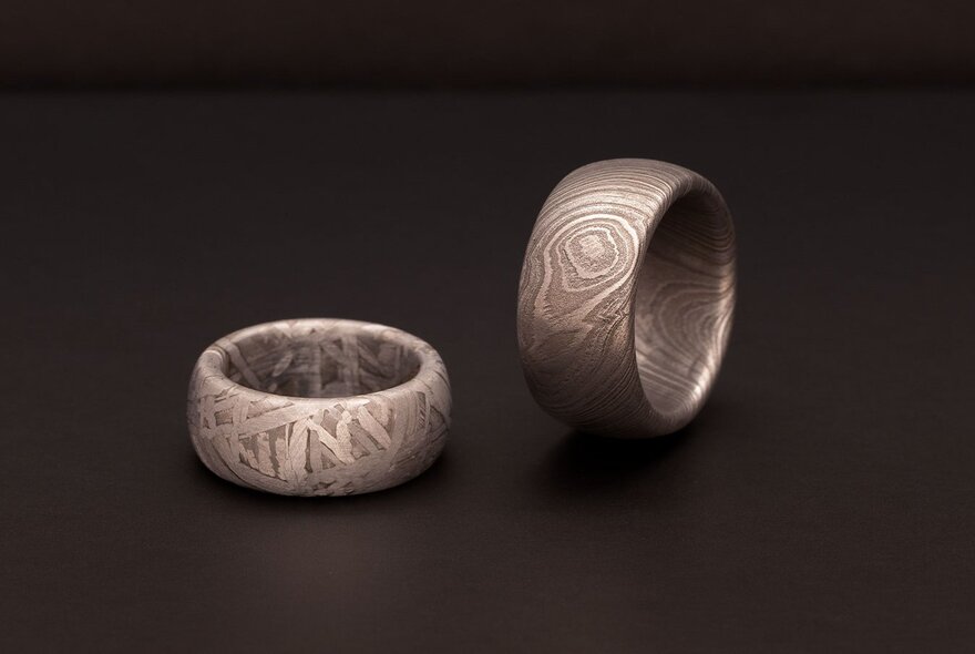 Two thick, patterned metal rings sit on a dark surface, with one lying flat and the other standing on its edge.