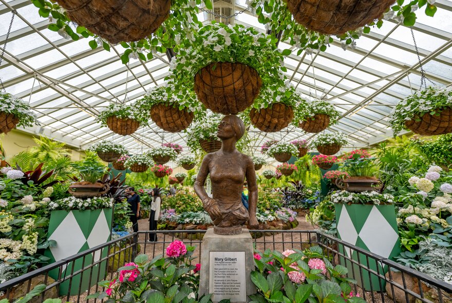 A statue of a woman surrounded by an abundance of plants, flowers and hanging pots in a greenhouse.