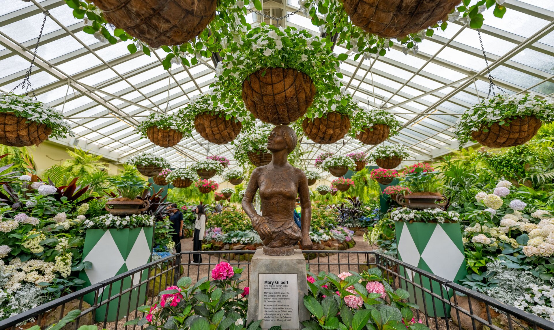 A statue of a woman surrounded by an abundance of plants, flowers and hanging pots in a greenhouse.