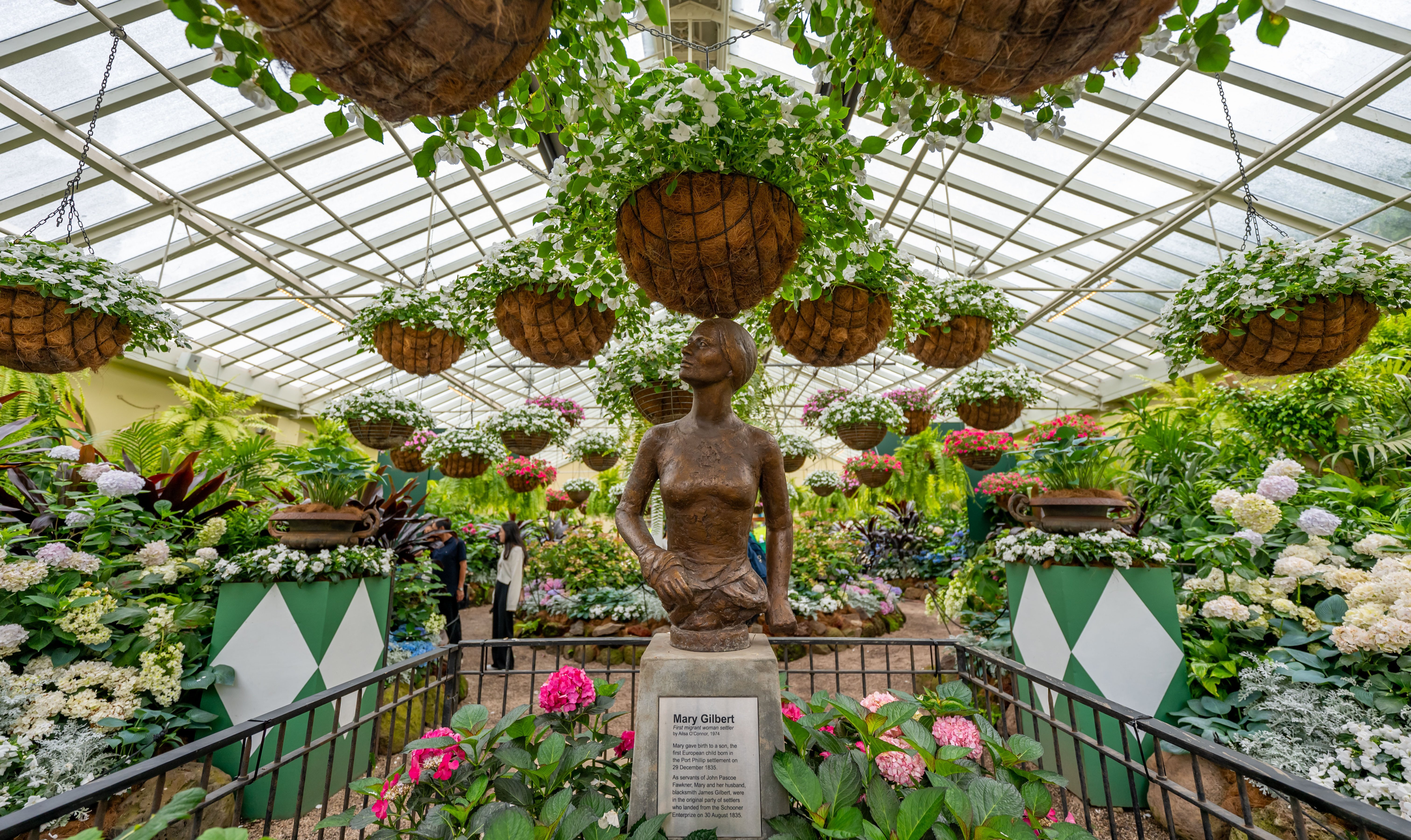 A statue of a woman surrounded by an abundance of plants, flowers and hanging pots in a greenhouse.