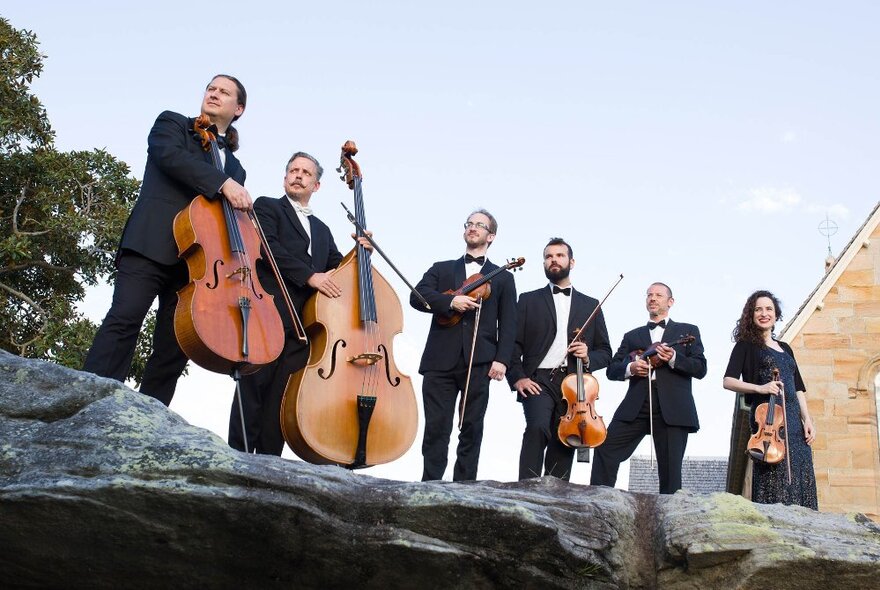 Looking up at members of Chamber Philharmonia Cologne holding their instruments and standing on the edge of a large rock. 