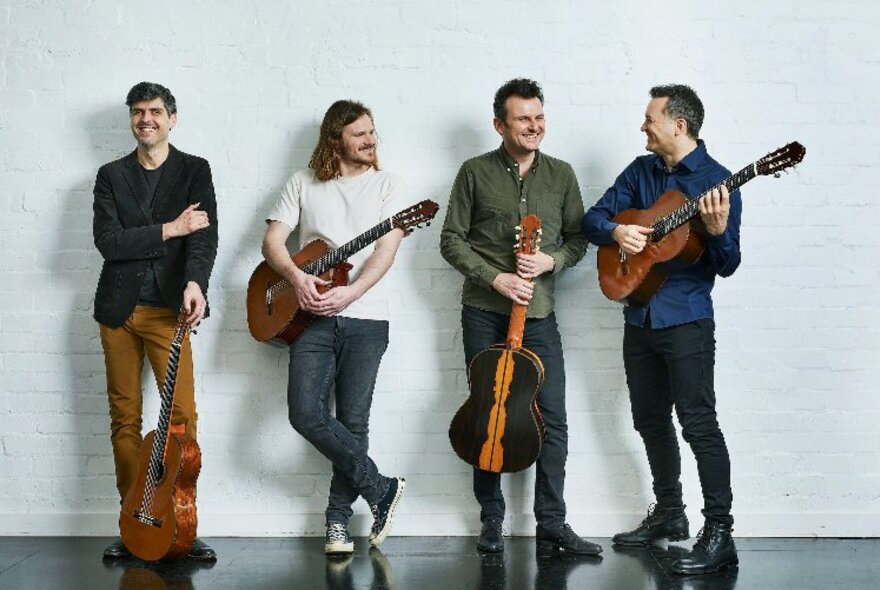 Four men standing against a white wall wearing casual clothes, smiling and interacting, and each holding a classical guitar in their hands.