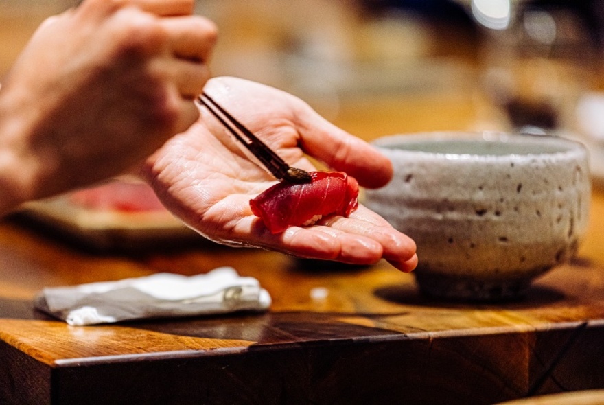 Hands holding a brush over a piece of sushi on a wooden table next to a pottery bowl.