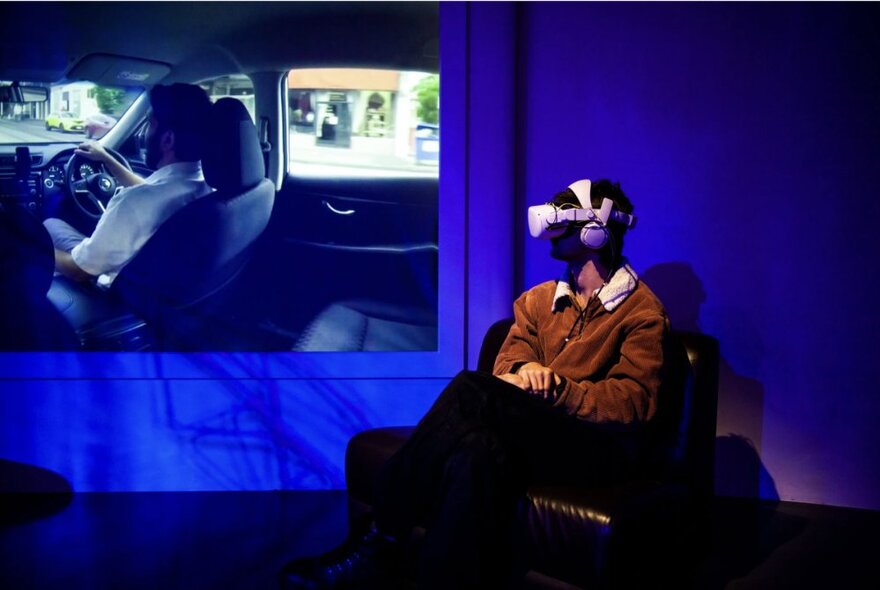 Person wearing a VR headset, seated in a chair with their head turned towards a projection on a wall to the side.