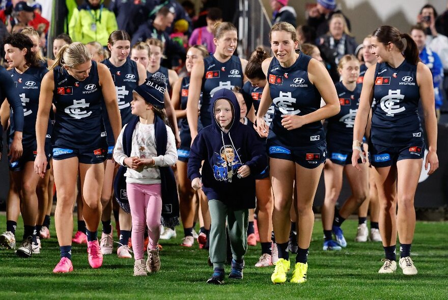 Carlton AFLW team members walking onto the field with child mascots.