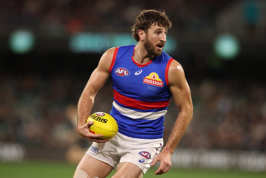 A Western Bulldogs AFL player running on the field while holding a yellow football in one hand.