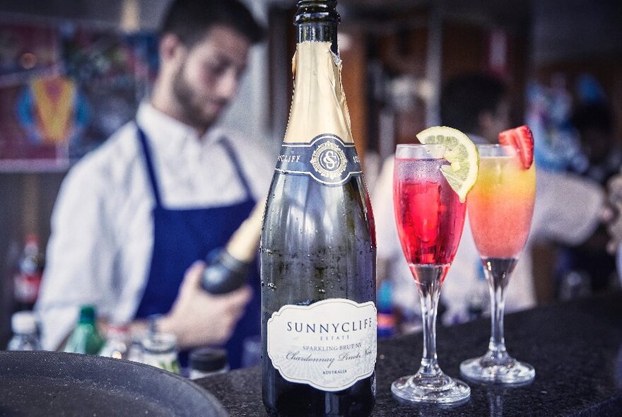 A bottle of champagne and several colourful cocktails with a bartender blurred in the background.