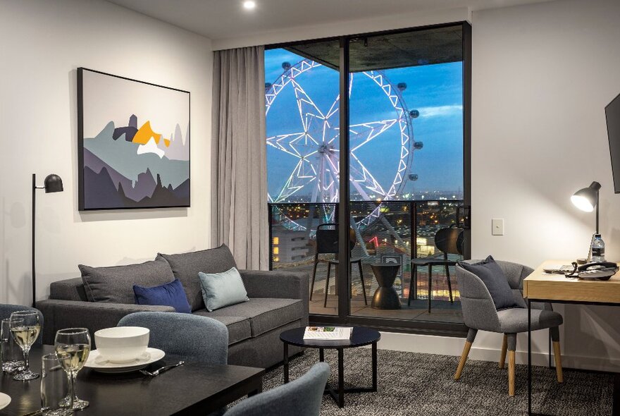 A room at Quest NewQuay with a sofa, a small dining table, a desk with chair and lamp and a view out the large window onto the Melbourne  SkyWheel.