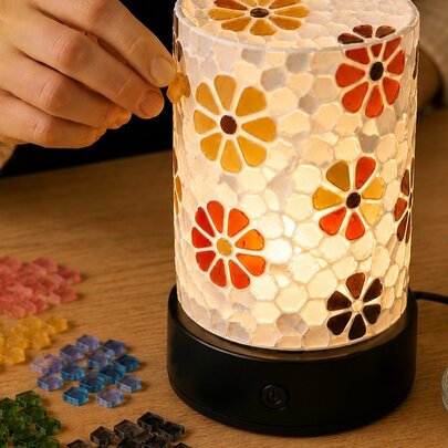 A cylindrical mosaic glass table lamp featuring a colourful orange and yellow daisy pattern, being made at a craft workshop.
