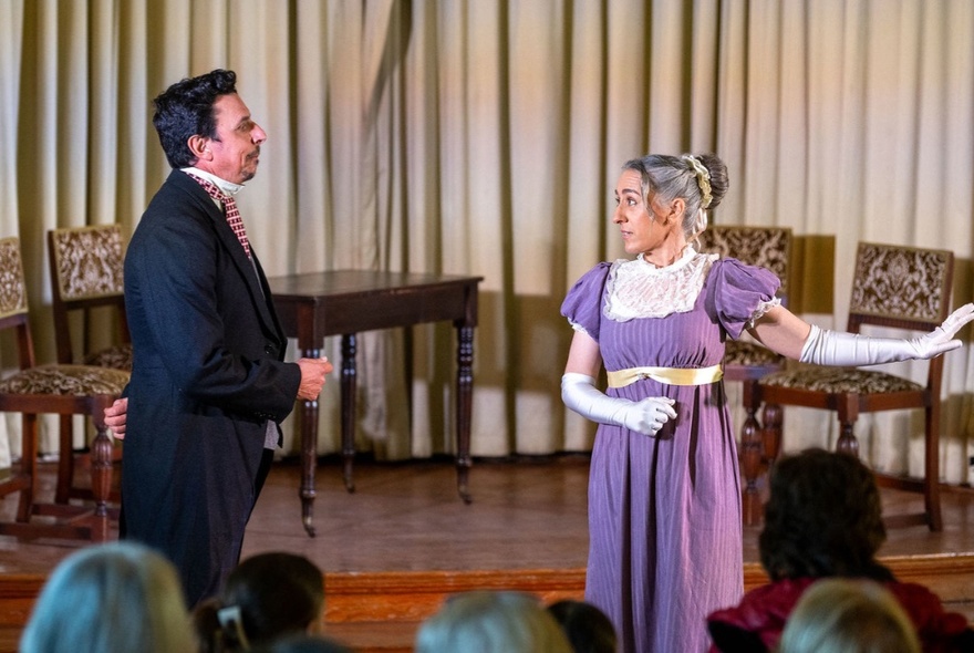 Two impro actors in Regency period costumes performing on a stage in front of a seated audience. 