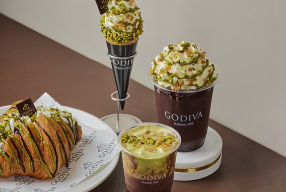 A selection of pistachio flavoured products on a table top, including a soft serve ice-cream, a croissant, a parfait and a chocolate drink.