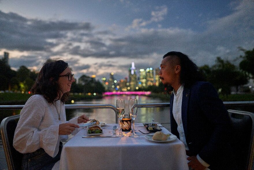 Two people seated opposite each other across a  dining table on a boat, with a view of the city skyline and river at dusk behind them.