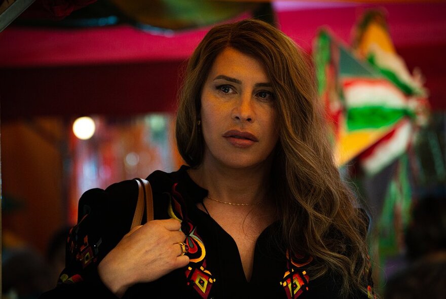 A still from the movie Emilia Pérez where a woman with long hair holds the straps of her handbag, in front of a colourful Mexican-style background.