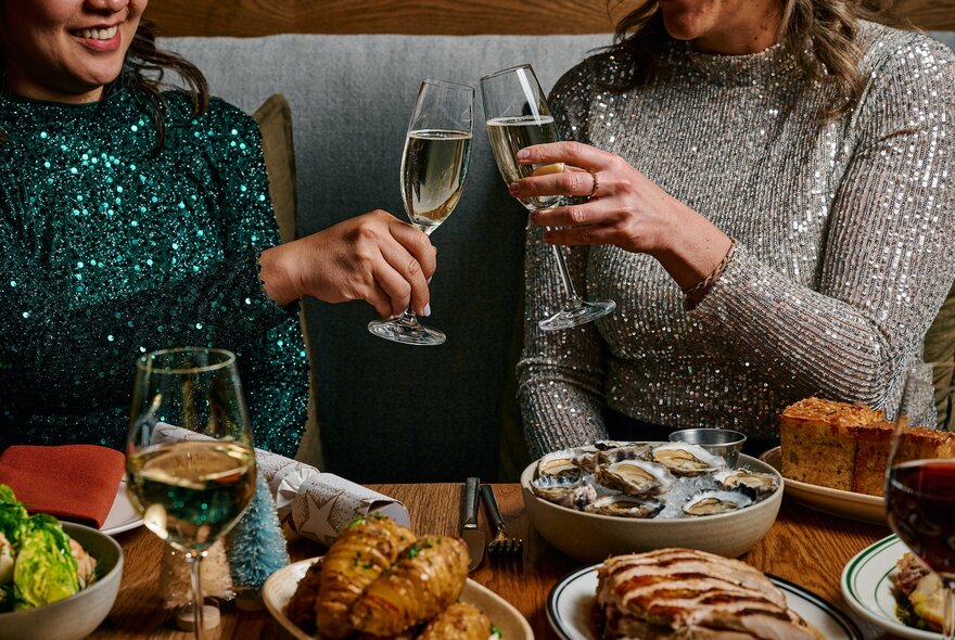 Two women wearing sparkly tops touching their wine glasses together in celebration, while seated at a table filled with Christmas food.
