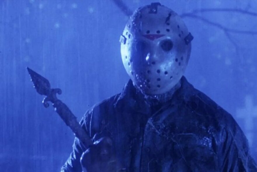 A still from a film showing a man in a perforated mask in the rain at night, holding a weapon.