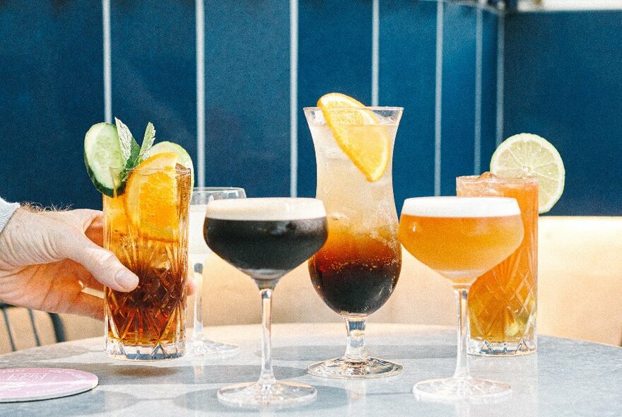 A hand placing a dark beverage in a tall glass on a table that contains four other beverages in assorted shaped glasses.