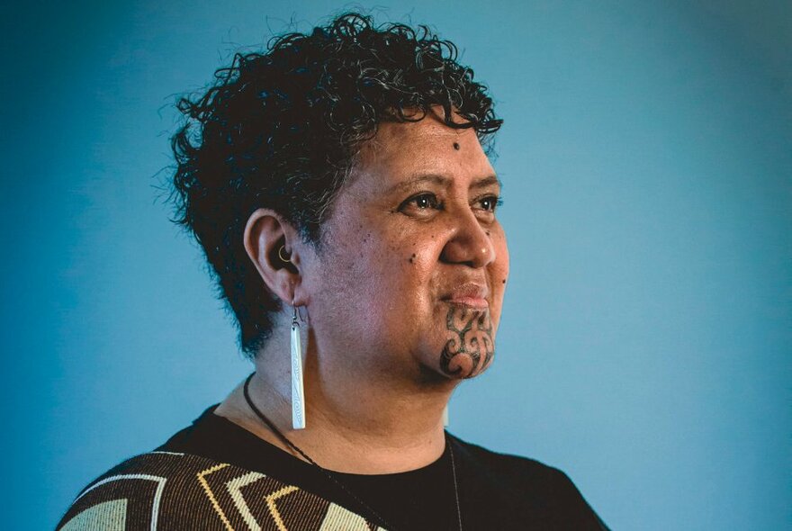 A Māori woman with a moko kauae (traditional chin tattoo) looking to her left and smiling proudly against a blue background. 