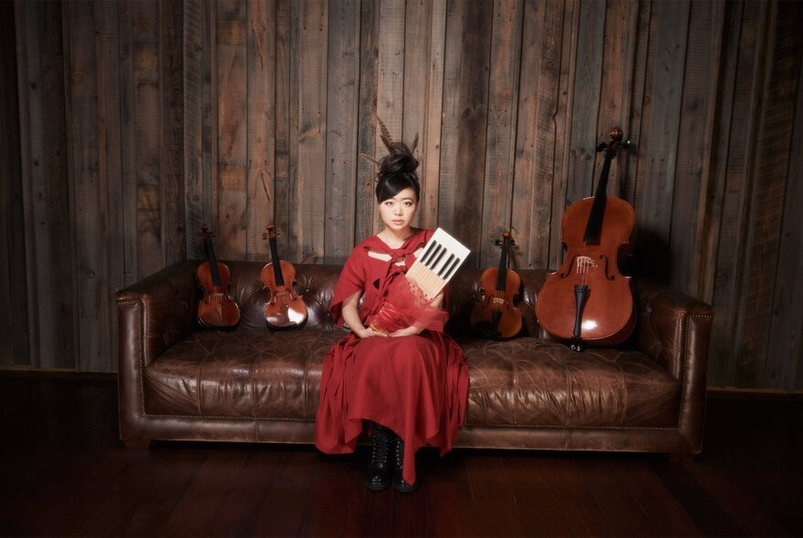Woman seated on a brown leather couch wearing a red gown, holding a keyboard instrument in red tulle.