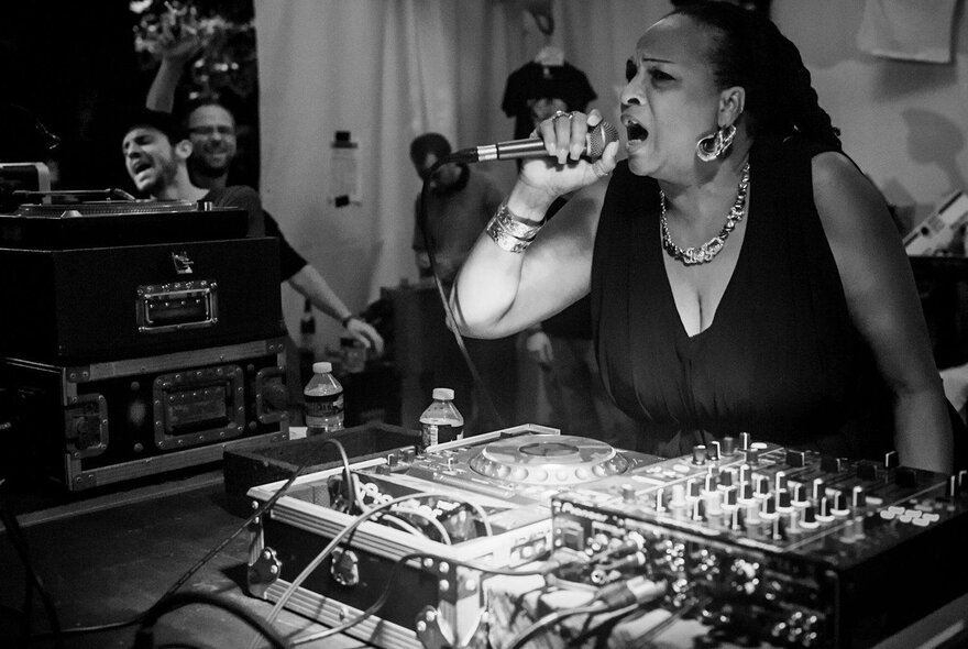 Black and white photograph of a musician singing into a microphone while leaning over a set of DJ decks.