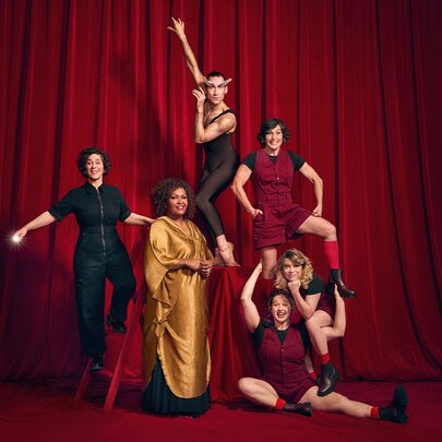 Six performers posing dramatically against a vibrant red stage curtain backdrop.