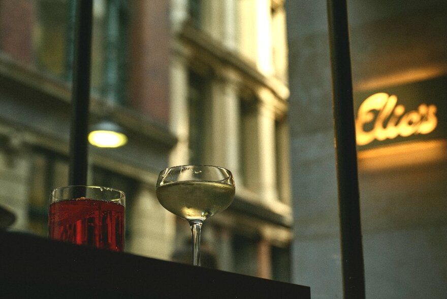 A champagne and negroni cocktail on a bartop, seen from inside a cafe looking out through the window to the buildings beyond, with a lit up sign that says 'Elio's'.