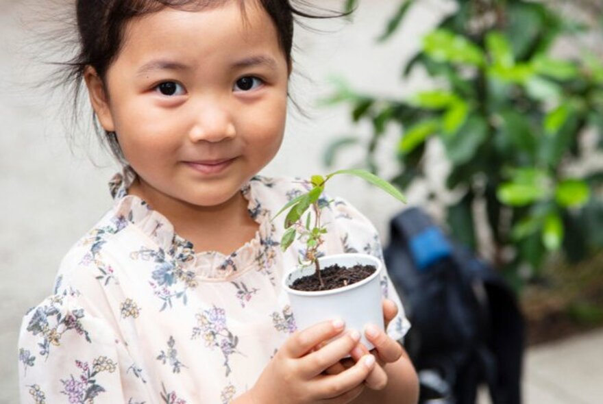 A child holding a tiny plant in a small white pot, plants in the background.