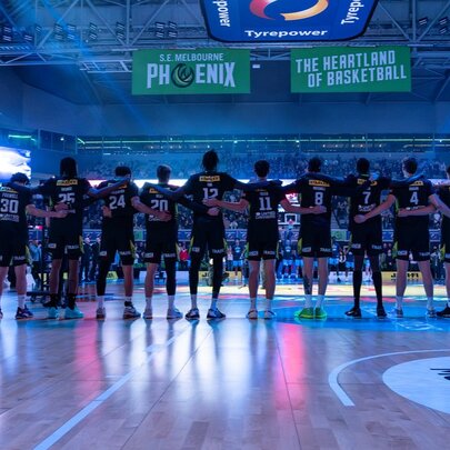 Back view of the South East Melbourne Phoenix basketball team, with linked arms, standing on a stadium court under basketball signage.