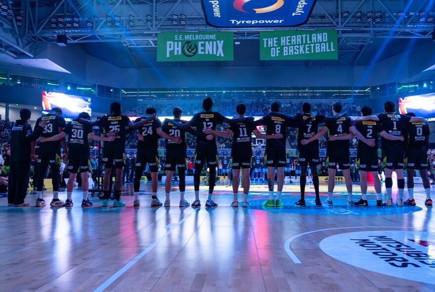 Back view of the South East Melbourne Phoenix basketball team, with linked arms, standing on a stadium court under basketball signage.